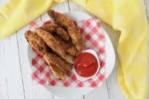 Overhead shot of finished chicken fingers in a white shallow bowl lined with red and white checkered picnic paper with a white dish of ketchup in the bowl on a white wooden surface with a yellow dish towel next to it