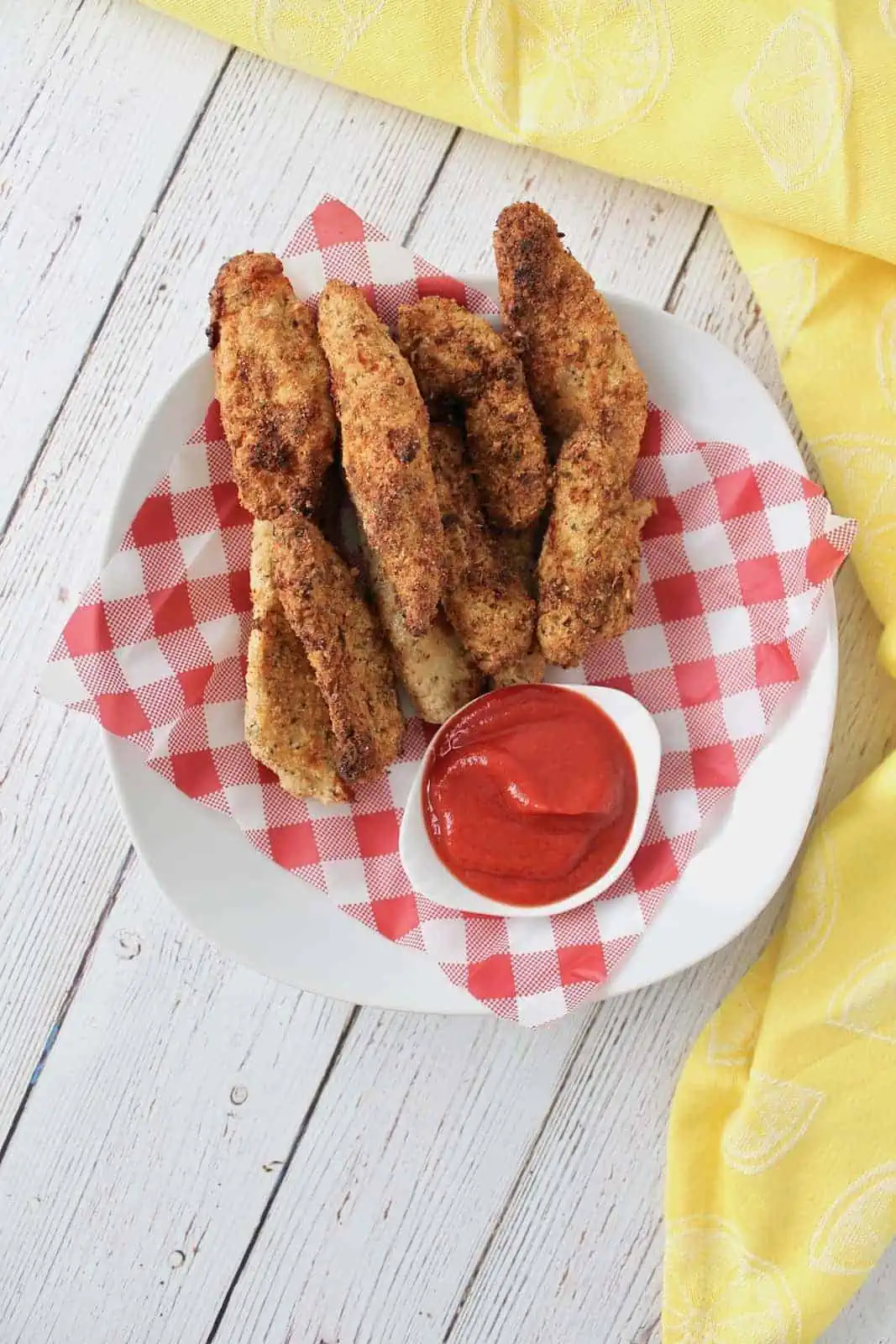 Overhead shot of finished chicken fingers in a white shallow bowl lined with red and white checkered picnic paper with a white dish of ketchup in the bowl on a white wooden surface with a yellow dish towel next to it