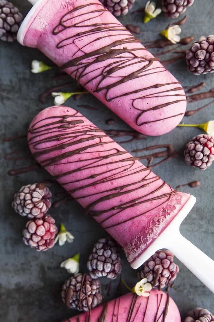 Two pink berry popsicles drizzled with chocolate surrounded by raspberries