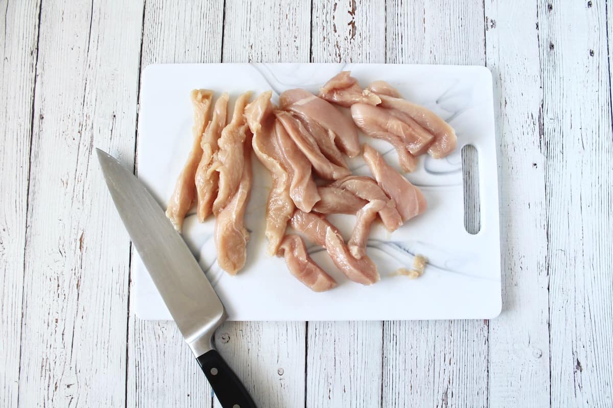 Overhead shot of sliced chicken breast on a white cutting board with a knife next to it on a white wooden surface
