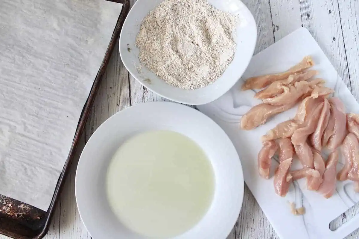 Overhead shot of raw chicken breast on a white cutting board next to a plate of oil, a bowl of flour mixture and a baking sheet lined with parchment paper