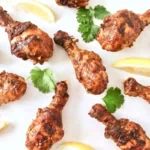 Cooked chicken tandoori on a white surface next to cilantro and sliced lemons