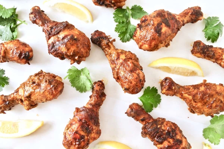 Cooked chicken tandoori on a white surface next to cilantro and sliced lemons