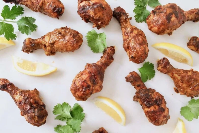 Cooked chicken tandoori on a white surface next to cilantro and sliced lemons