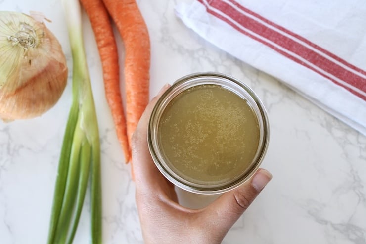 Overhead view of hand holding a jar of bone broth with carrots and an onion on the table