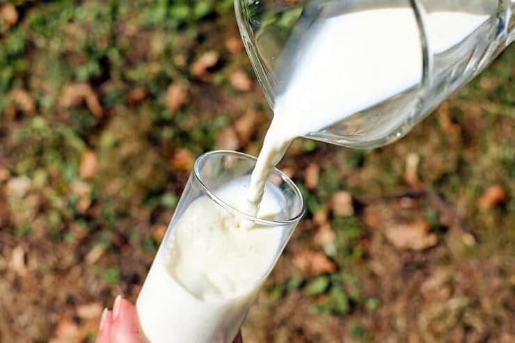 A hand holding a glass of milk and pouring milk into it from a jug