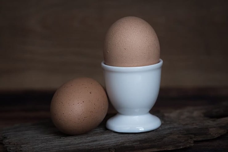 Two eggs one in an egg cup and one beside it sitting on a wooden surface