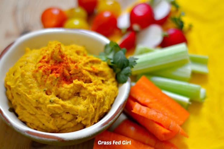 Small white bowl of orange colored pumpkin hummus surrounded by chopped vegetables