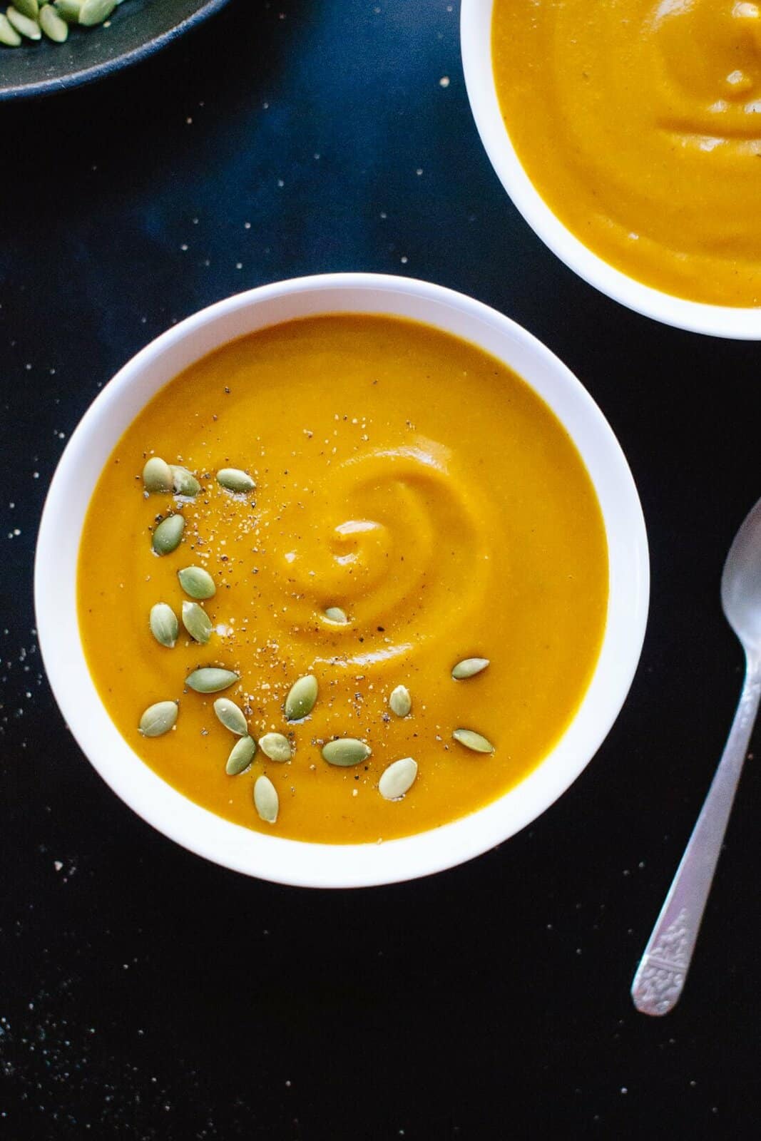 overhead shot of white bowl filled with orange colored pumpkin soup with pumpkin seeds sprinkled on top on a black surface