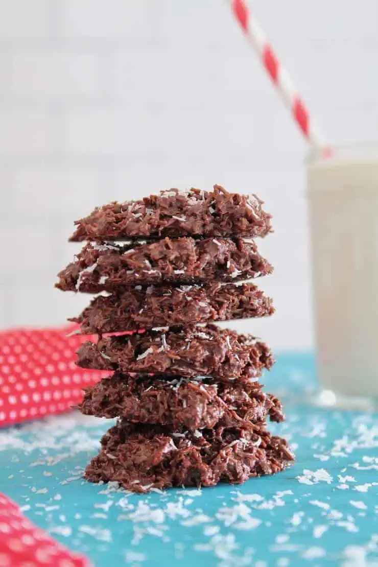 stacked coconut haystack cookies on a blue surface next to a glass of milk and a red and white polka dot napkin