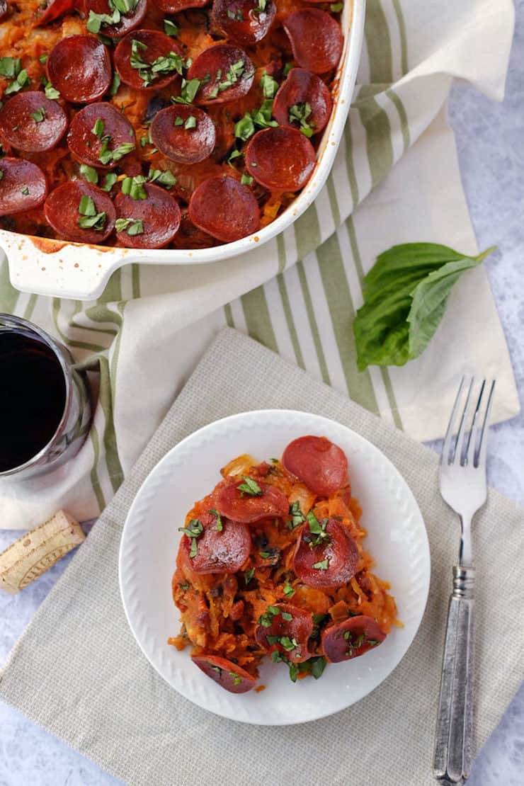 White casserole dish with pepperoni topped lasagna and a serving on a white plate next to it