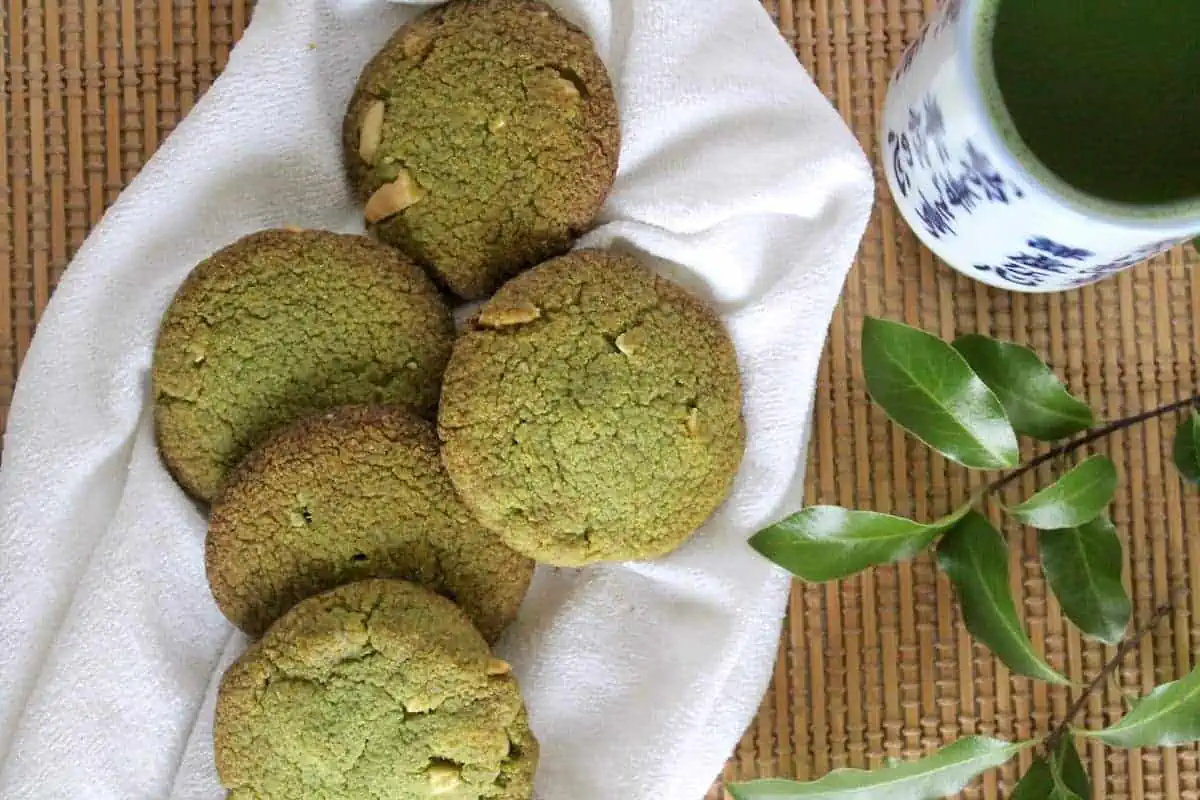Five matcha green tea cookies on a white surface with a small mug of green matcha tea on the side