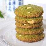 Five stacked matcha green tea cookies on a white plate with a cup of green tea and green leaves in the background