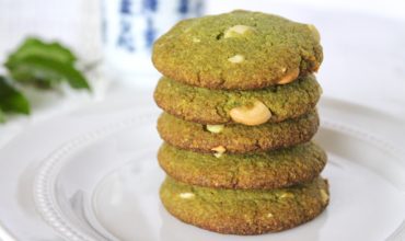 Five stacked matcha green tea cookies on a white plate with a cup of green tea and green leaves in the background
