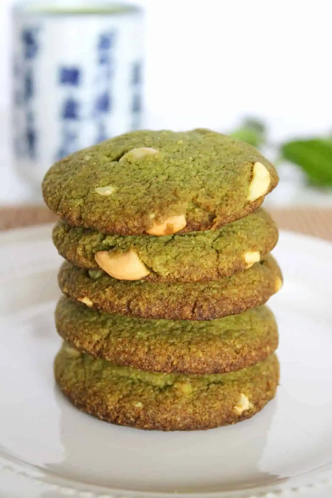 Close up of five stacked matcha green tea cookies on a white plate with a mug of green tea and green leaves in the background