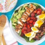 An overhead of a cobb salad sitting on a blue surface with salad servers at the side