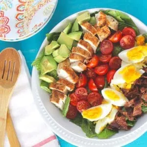 An overhead of a cobb salad sitting on a blue surface with salad servers at the side