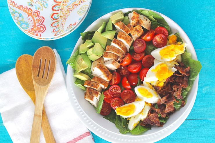 An overhead of a cobb salad sitting on a blue surface with salad servers at the side