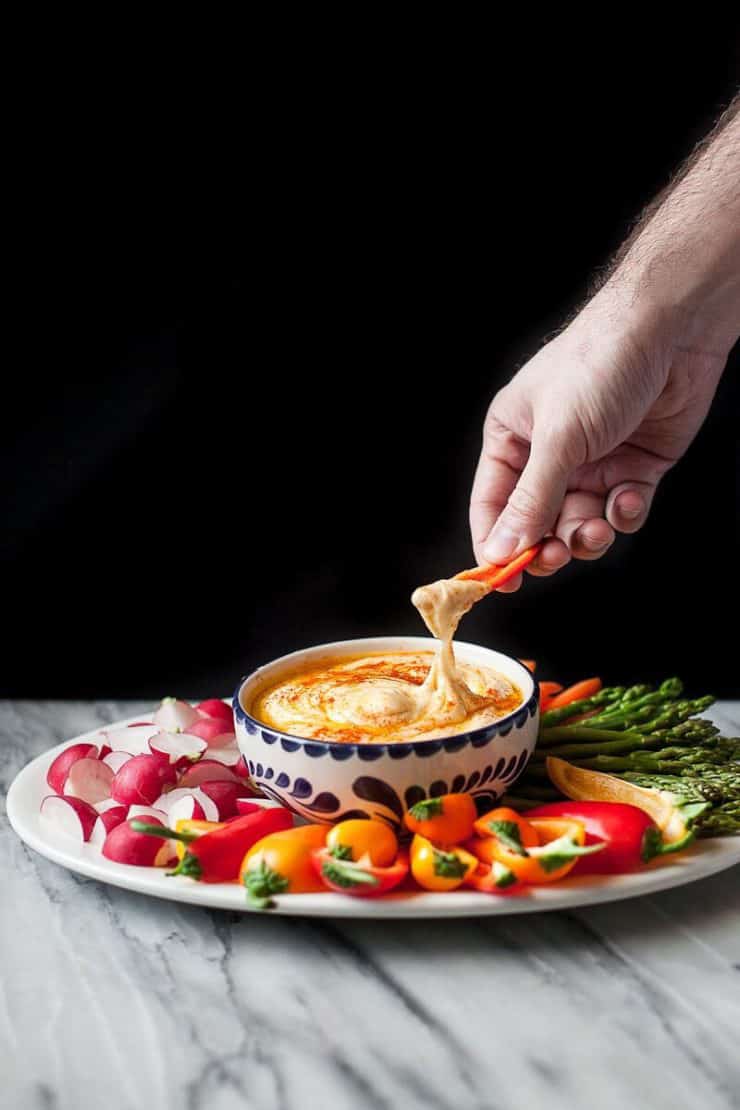 A bowl of potato hummus with vegetables around it and a hand dipping a carrot into it
