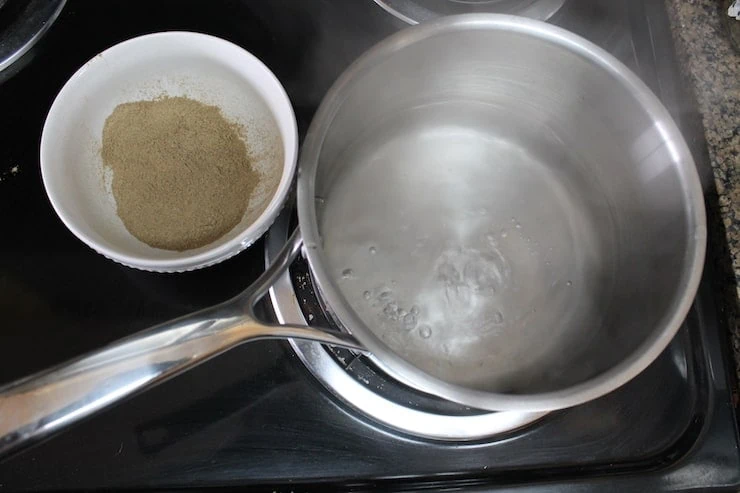 stainless steel pot on the stove with boiling water inside it beside bowl of powdered essiac tea