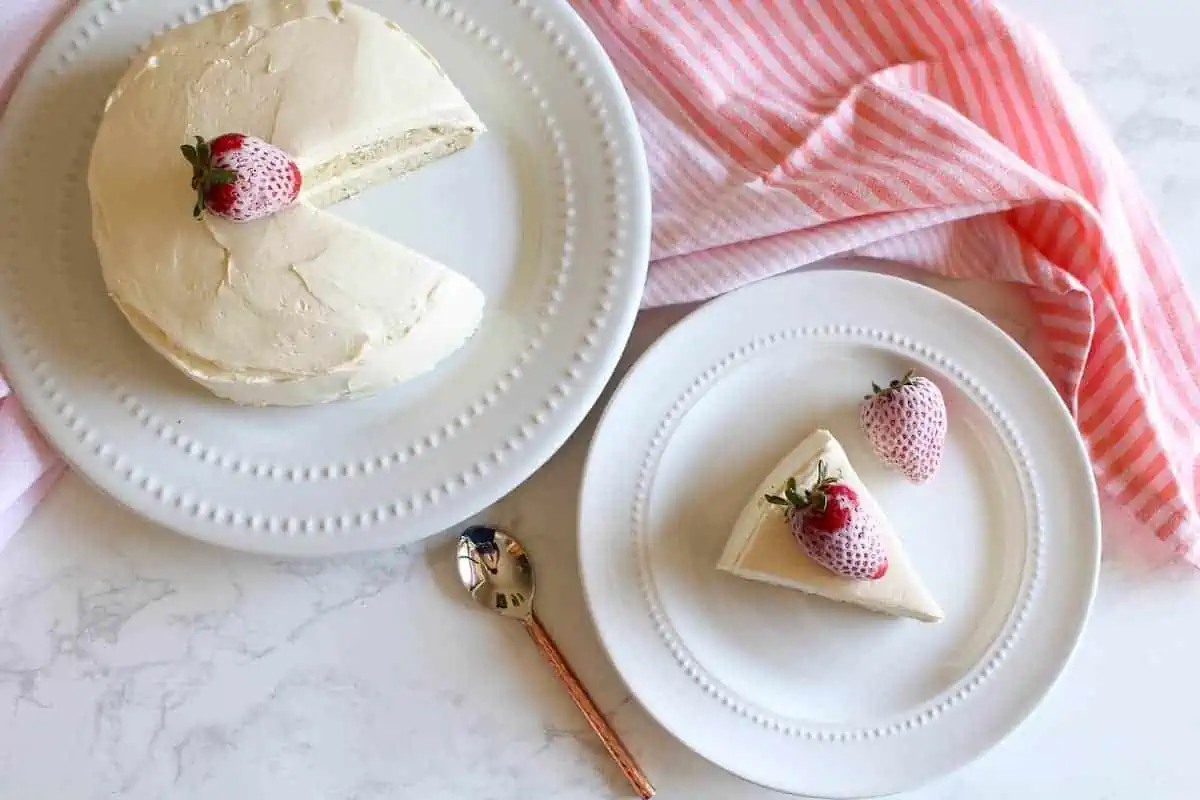 Overhead view of white frosted coconut flour cake with strawberries on top on white plate with slice of cake on smaller white plate next to it