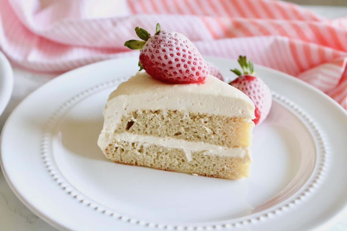 Slice of white frosted coconut flour layer cake on white plate with strawberry on top next to pink striped dish towel