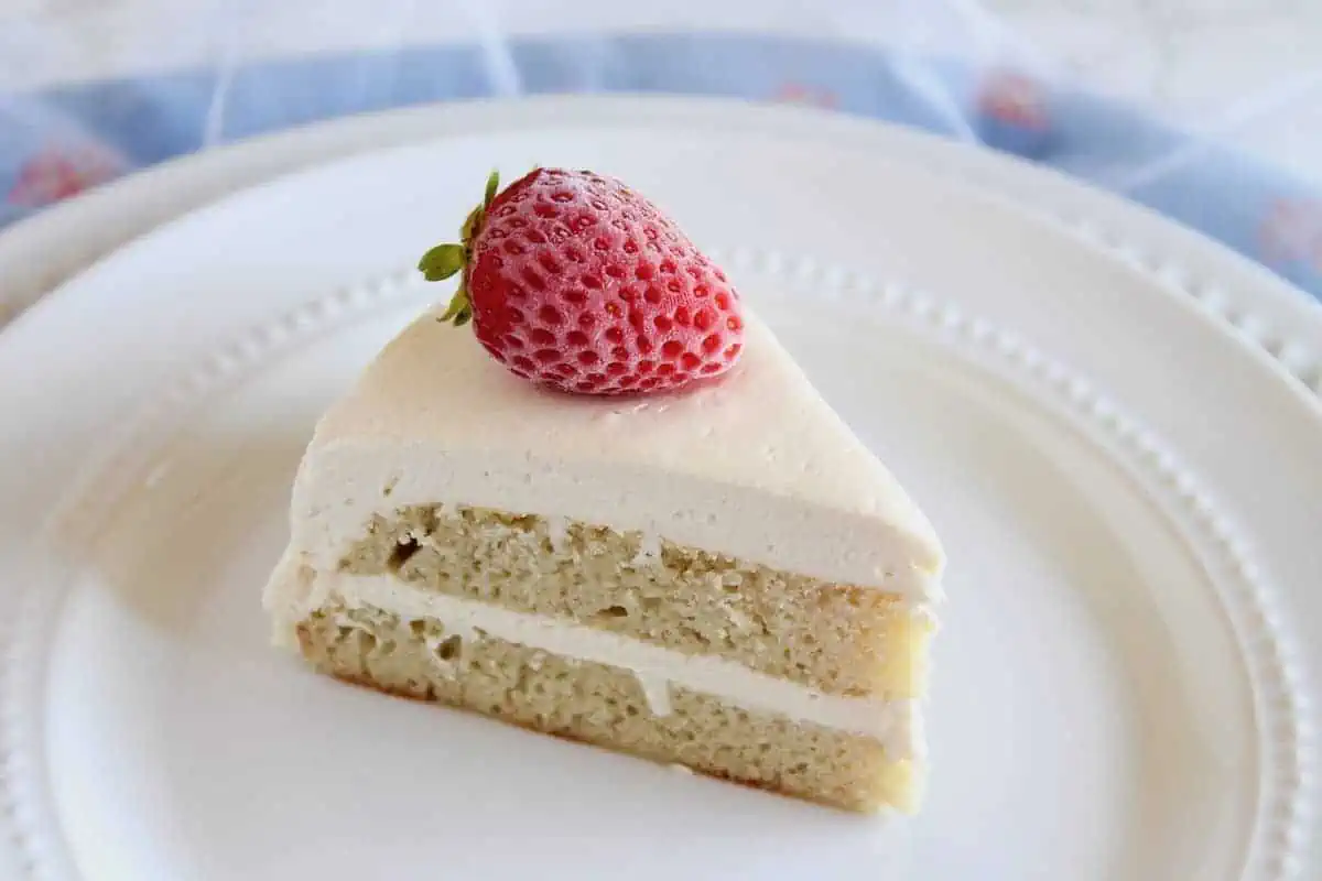 Slice of white frosted coconut flour layer cake on white plate with strawberry on top next to blue cloth
