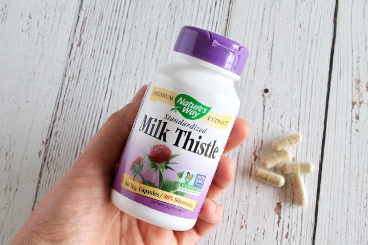 Hand holding bottle of milk thistle with tan capsules in background on white wooden table