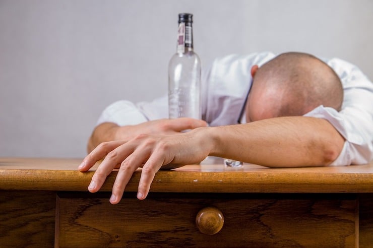 Man with head down on desk holding a bottle of alcohol