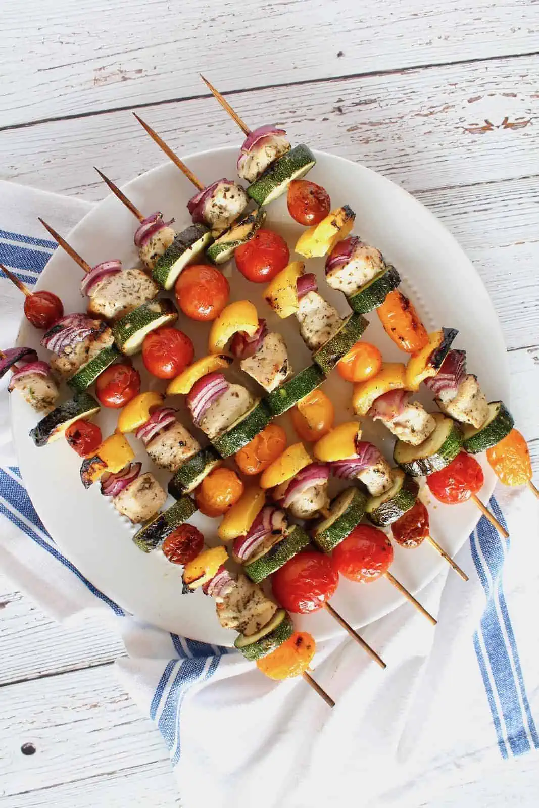 overhead view of cooked chicken kabobs on a white plate with white cloth with blue stripe underneath