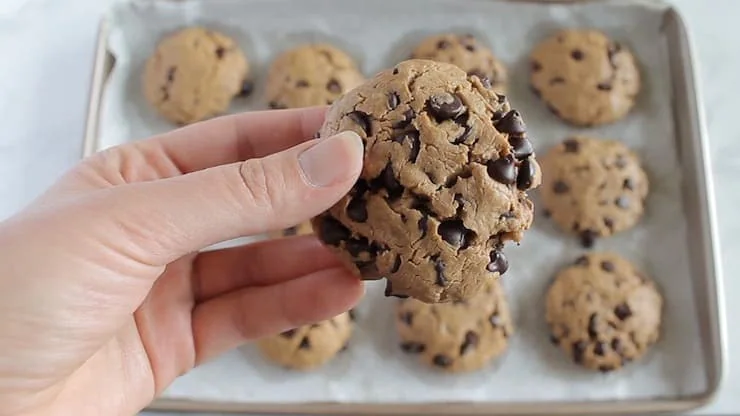 Hand holding one keto chocolate chip cookie with baking sheet with cookies in the background