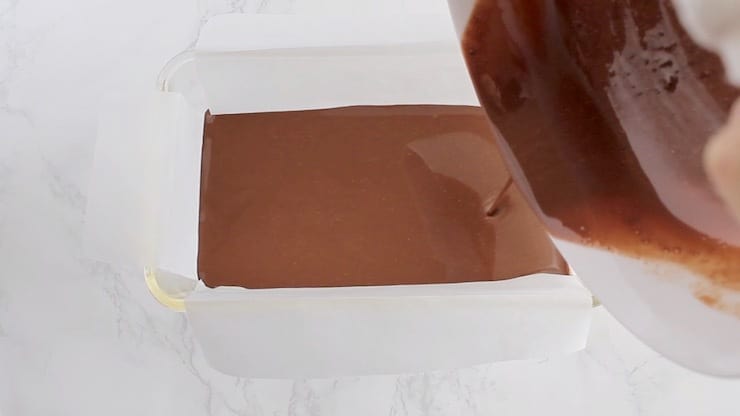Keto fudge melted ingredients being poured into a glass loaf pan lined with white parchment paper