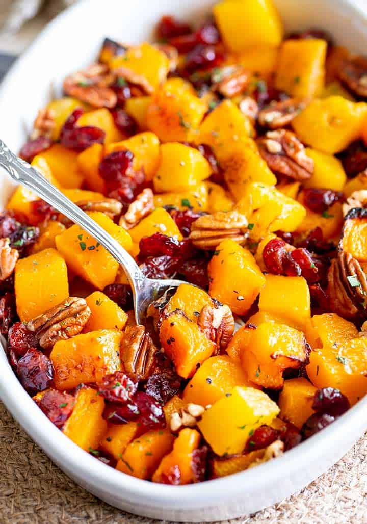 white dish filled with roasted pumpkin with cranberries and pecans with a spoon in it