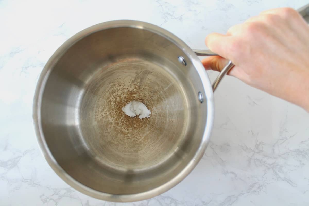 stainless steel pot with a small amount of coconut oil in it with a hand grasping the handle