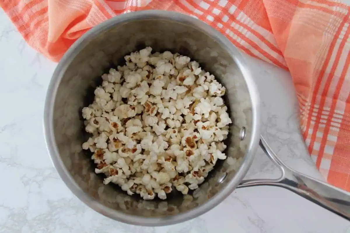 Stainless steel pot with popped popcorn in it on a white marble surface next to a red and white plaid dish cloth