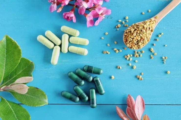 Wooden spoon full of bee pollen next to light and dark green vitamin capsules on a blue wooden surface