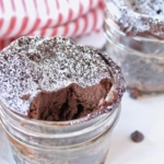 2 jars of finished chocolate keto mug cakes one with a bite taken out of it on a white surface sprinkled with chocolate chips next to a red and white striped dish towel