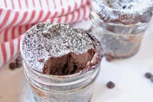 2 jars of finished chocolate keto mug cakes one with a bite taken out of it on a white surface sprinkled with chocolate chips next to a red and white striped dish towel