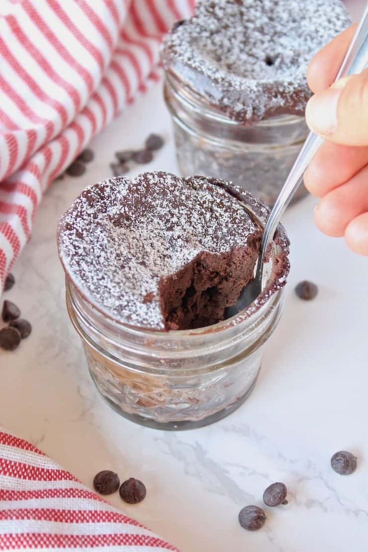 2 jars of finished chocolate keto mug cakes one with a spoon taking a piece out of it on a white surface sprinkled with chocolate chips next to a red and white striped dish towel