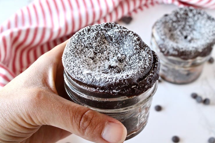 A hand holding a finished jar of chocolate keto mug cake over a white surface sprinkled with chocolate chips and a red and white striped dish towel