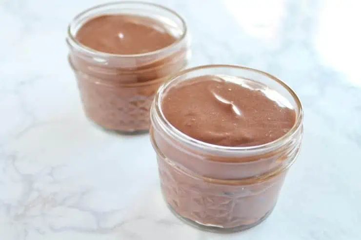 Uncooked chocolate keto mug cake batter in 2 small jars on a white marble surface