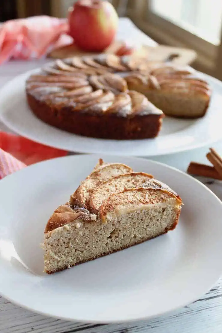 Slice of apple cake on white plate with entire cake in background next to cinnamon sticks and red and white plaid dish towel