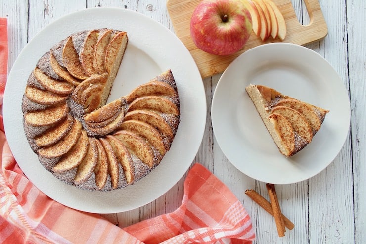 Overhead view of apple cake on a white plate with a slice out of it next to smal white plate with slice of apple cake on it