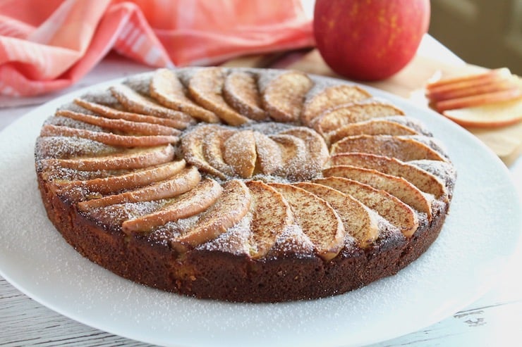 Whole apple cake with arranged apple slices on top on a white plate with an apple in the background next to red and white plaid dish towel