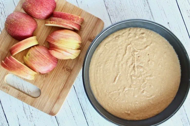 Uncooked apple cake batter in metal cake pan next to wooden cutting board with sliced apples on it on a white wooden table