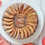 Overhead view of whole apple cake with arranged apple slices on top on a white plate with an apple in the background next to red and white plaid dish towel