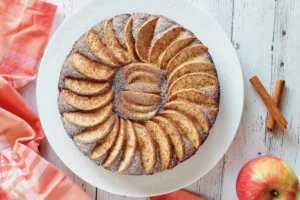 Overhead view of whole apple cake with arranged apple slices on top on a white plate with an apple in the background next to red and white plaid dish towel
