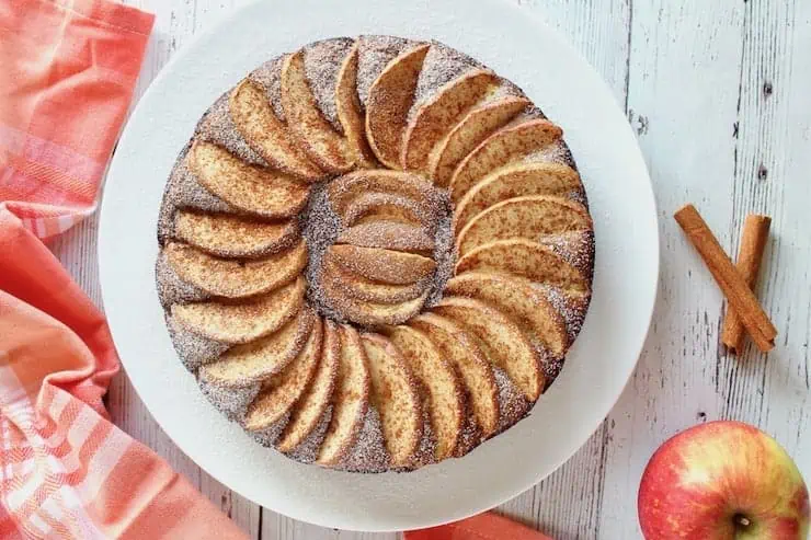 Overhead view of whole apple cake with arranged apple slices on top on a white plate with an apple in the background next to red and white plaid dish towel