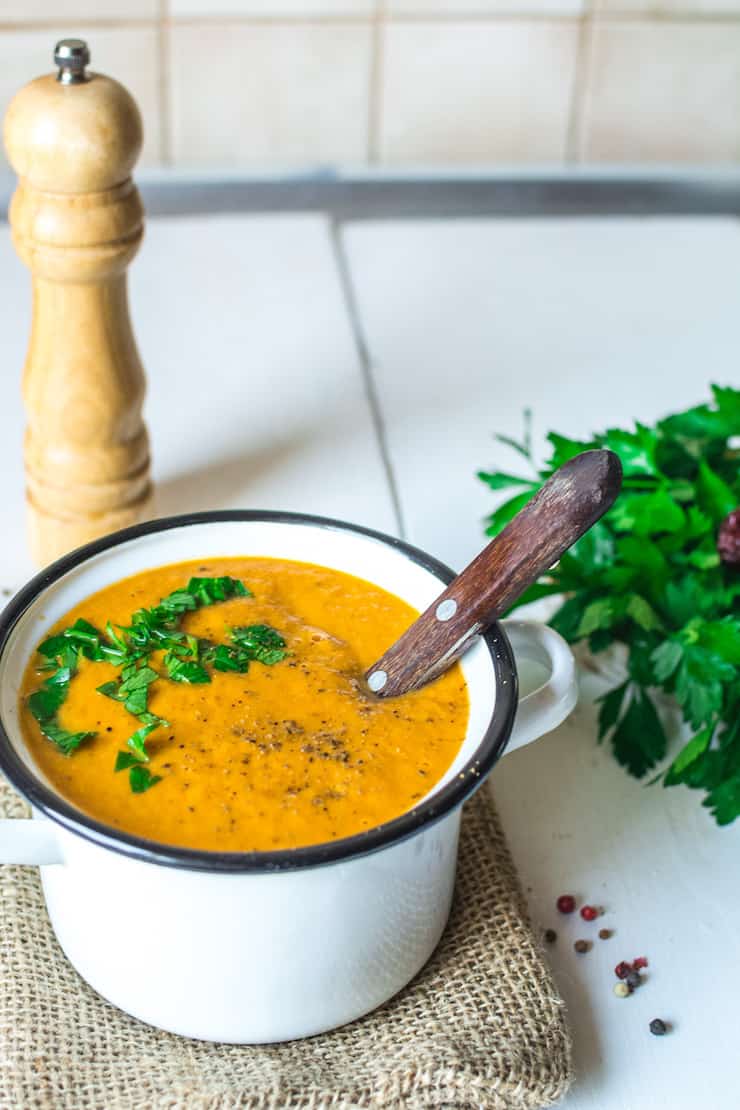 Creamy orange roasted vegetable soup topped with parsley in a white bowl on a burlap towel on a white wooden table next to a bunch of parsley and a pepper grinder