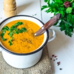 Creamy orange roasted vegetable soup topped with parsley in a white bowl on a burlap towel on a white wooden table next to a bunch of parsley and a pepper grinder